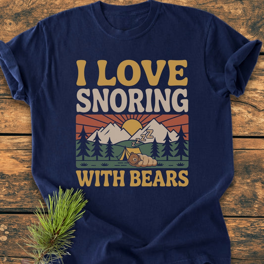 Navy-blue I Love Snoring With Bears - Vintage Wash Tee featuring a retro camping graphic with a cartoon bear, tent, and sunrise. Displayed on rustic wooden surface with a pine sprig accent.