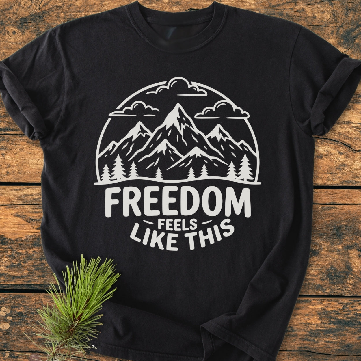 Freedom Feels Like This - Vintage Wash Tee features a stylized mountain scene with FREEDOM text. Displayed on rustic wood, the black tee is accompanied by a pine sprig, showcasing cozy, durable design.