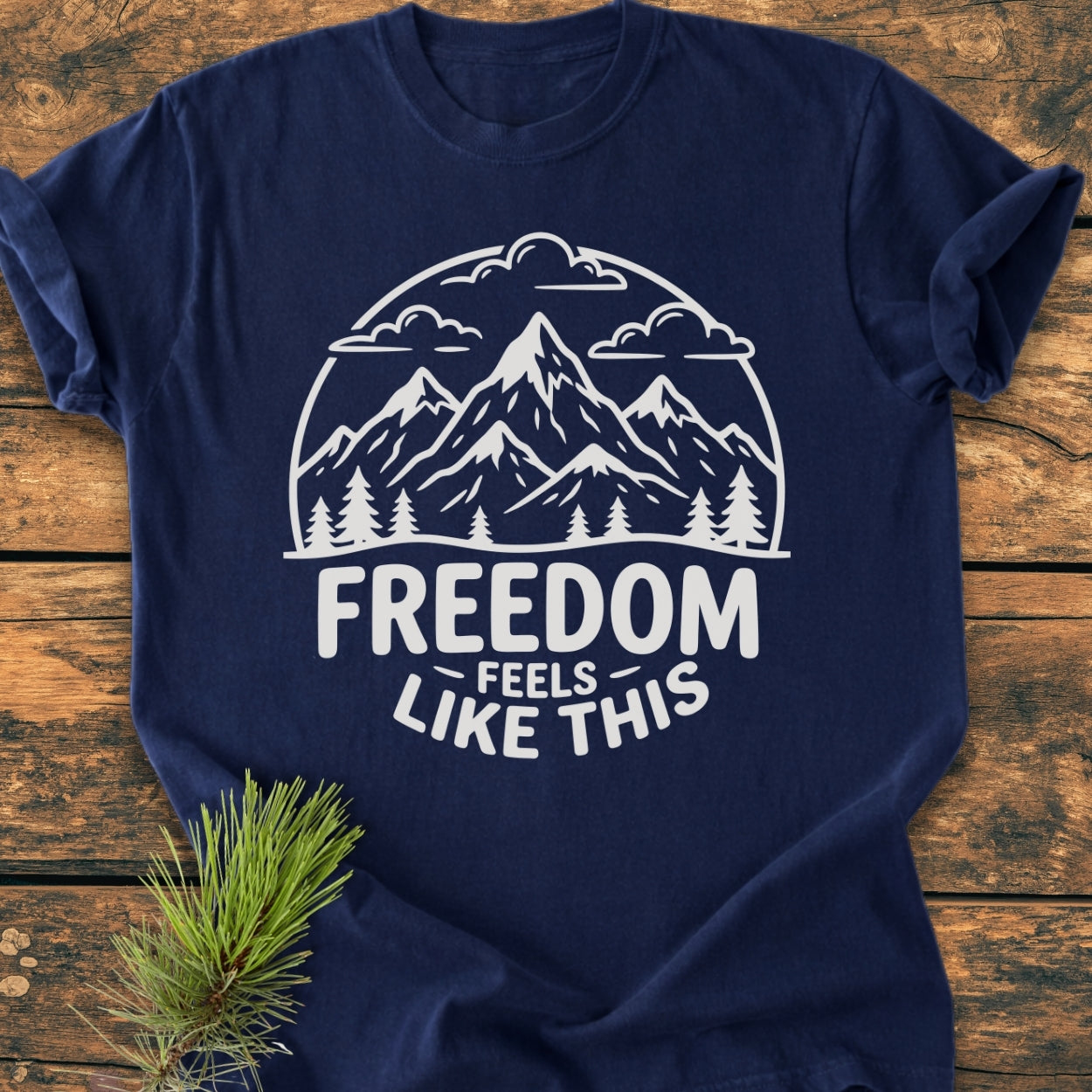 Freedom Feels Like This - Vintage Wash Tee features a stylized mountain scene with slogan on navy-blue fabric, laid on rustic wood. A pine sprig complements this Comfort Colors garment-dyed T-shirt.