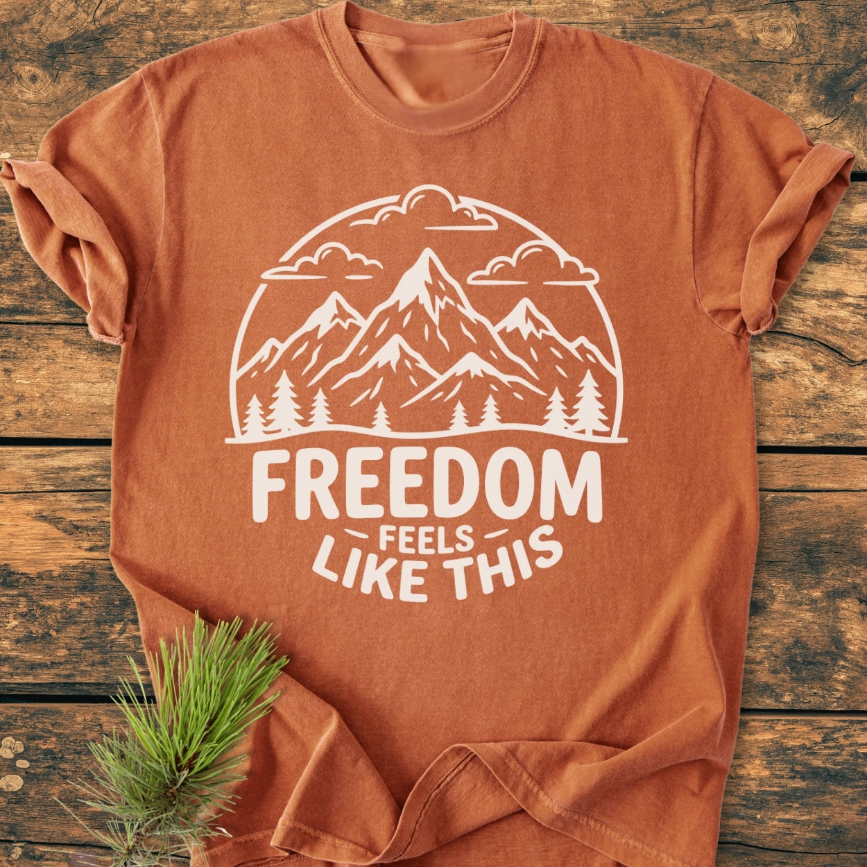 Freedom Feels Like This - Vintage Wash Tee: Rust-orange T-shirt with white mountain and pine design, slogan underneath. Laid flat, pine sprig beside. Made of 100% ring-spun cotton, relaxed fit.