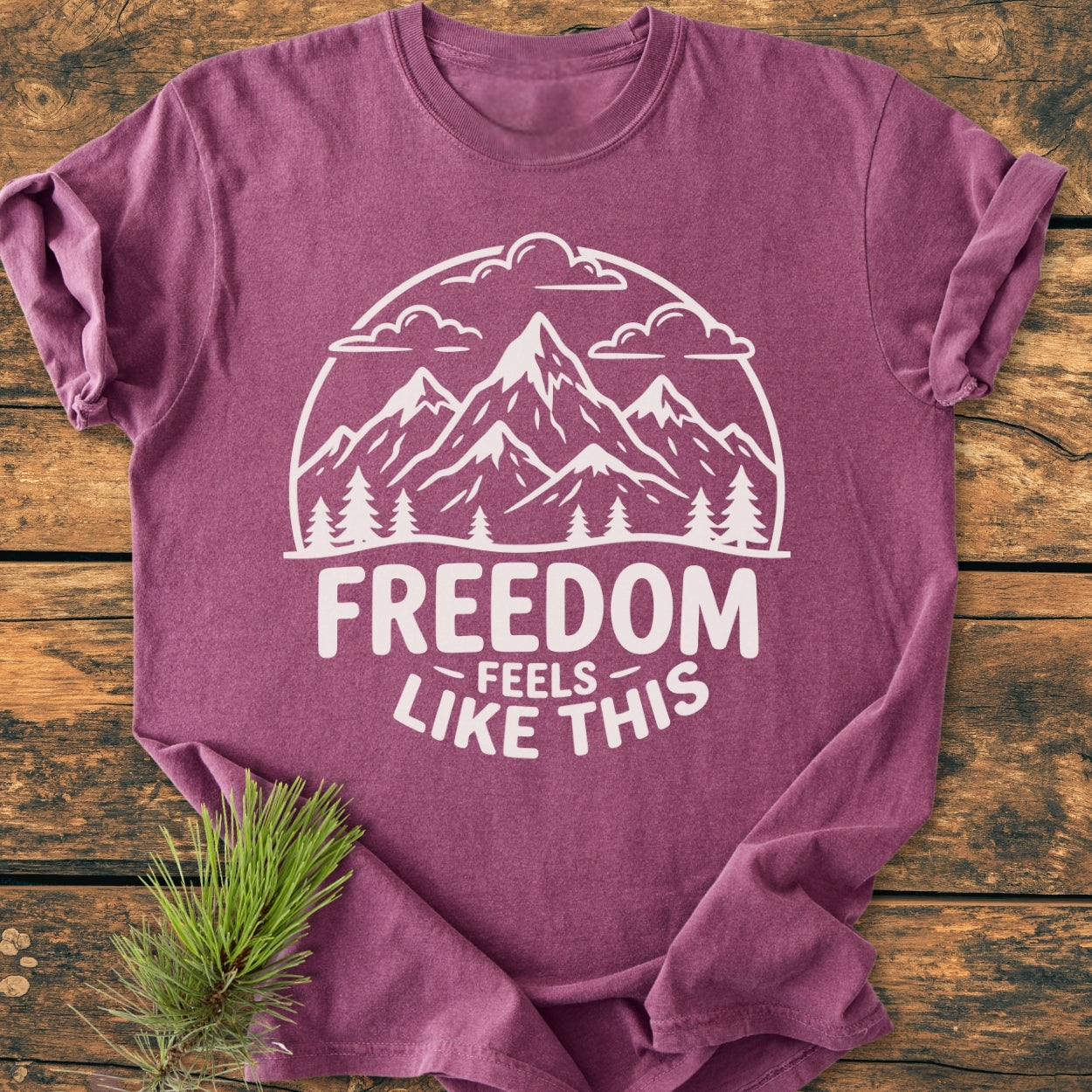 Freedom Feels Like This - Vintage Wash Tee displays a mountain scene and slogan, laid on wood with a pine branch. Made of soft-washed, ring-spun cotton, offering comfort and durability.