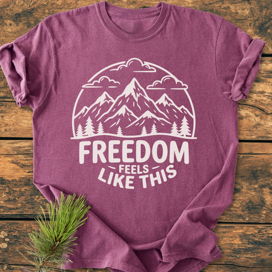 Freedom Feels Like This - Vintage Wash Tee displays a mountain scene and slogan, laid on wood with a pine branch. Made of soft-washed, ring-spun cotton, offering comfort and durability.