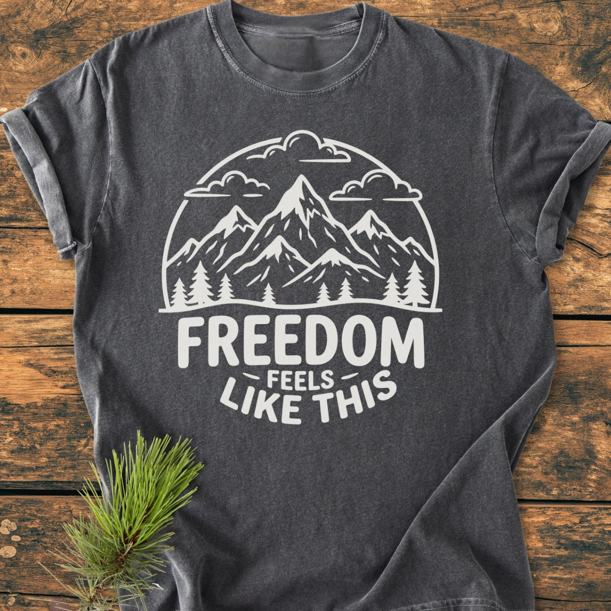 Freedom Feels Like This - Vintage Wash Tee: Charcoal-gray T-shirt with white mountain and tree graphic, bold text beneath. Laid on wood surface with pine sprig, emphasizing comfort and durability.