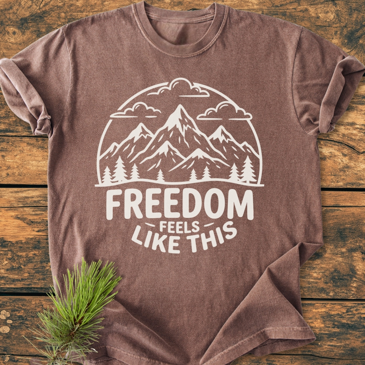 Freedom Feels Like This - Vintage Wash Tee displayed on wood, featuring a circular mountain scene with clouds and trees, slogan beneath. Nearby, a pine sprig enhances the rustic presentation.