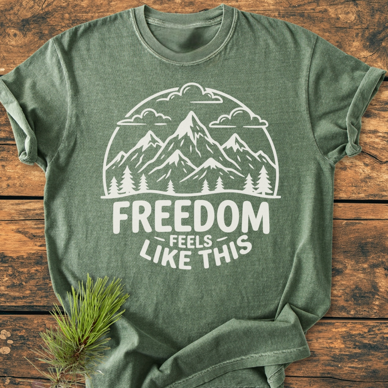 Freedom Feels Like This - Vintage Wash Tee features a green heathered fabric with a white outdoors graphic and slogan. Laid flat on wood, it embodies comfort and durability with its relaxed, pre-shrunk fit.