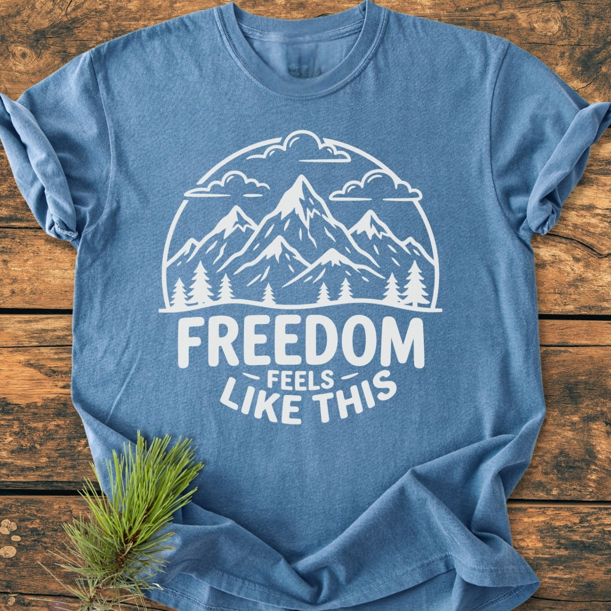 Heather-blue Freedom Feels Like This - Vintage Wash Tee with a white mountain graphic and text, displayed flat on wood beside a pine branch, highlighting its relaxed fit and cozy texture.