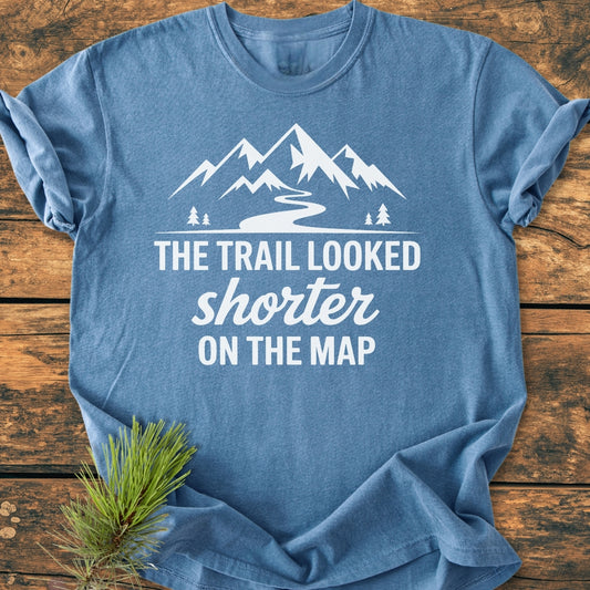 The Trail Looked Shorter On The Map - Vintage Wash Tee displays a stylized mountain range and slogan on soft, heather-blue fabric, laid flat with pine needles beside it.