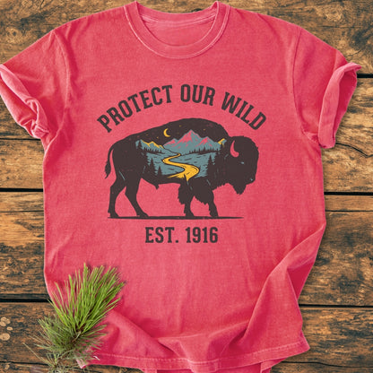 Protect Our Wild - Vintage Wash Tee features a buffalo silhouette with a mountain scene. Text reads 'PROTECT OUR WILD' and 'EST. 1916.' Laid on wood, showcasing its cozy, relaxed fit.