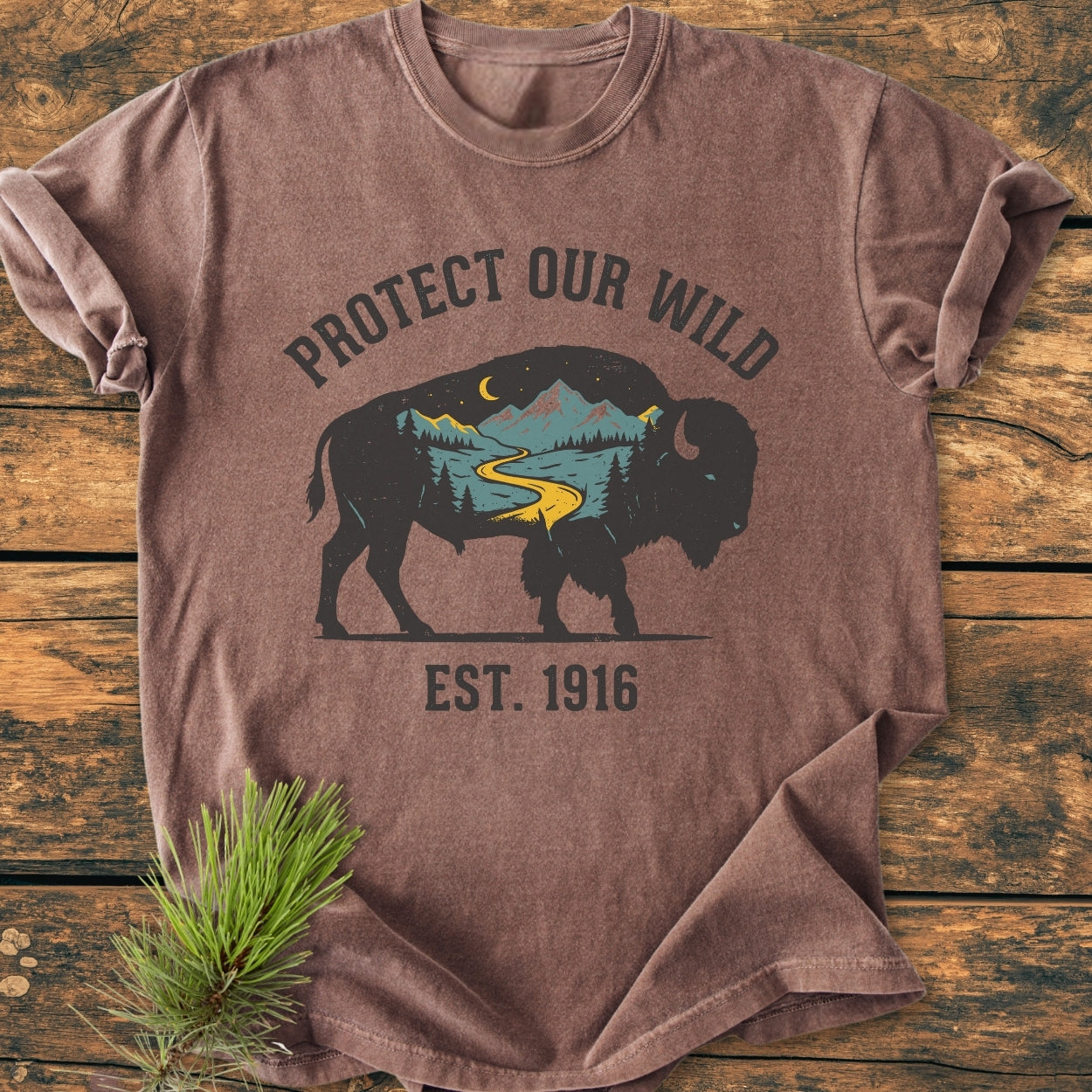 Protect Our Wild - Vintage Wash Tee featuring a bison silhouette with a nature scene and slogan. Laid on wood with a pine sprig, showcasing garment-dyed, relaxed fit, durable stitching, and pre-shrunk cotton.