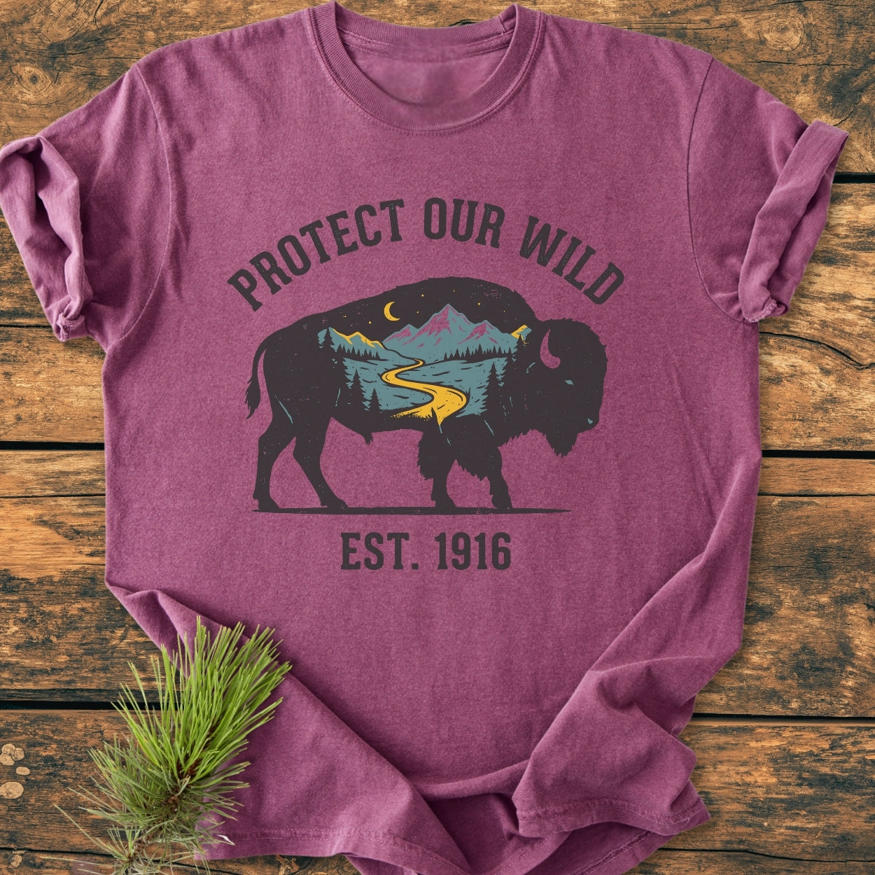 Protect Our Wild - Vintage Wash Tee featuring a bison silhouette with a mountain scene, crescent moon, and PROTECT OUR WILD text, laid on wood with a pine sprig nearby.