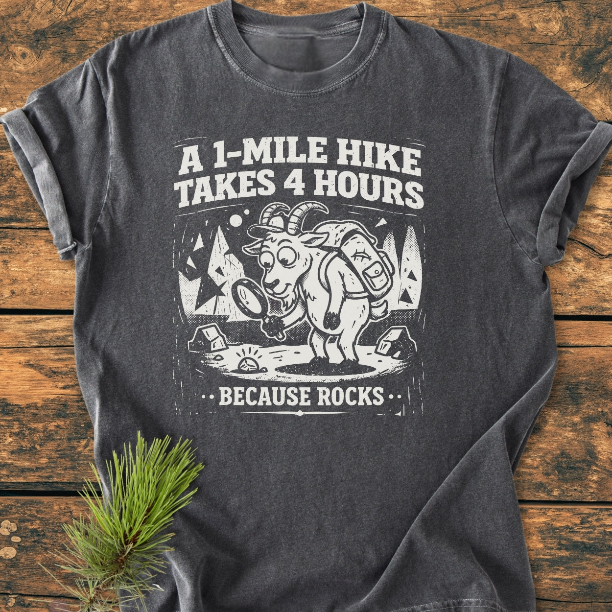 Because Rocks - Vintage Wash Tee featuring a cartoon goat with a backpack and magnifying glass against a forest, humorously captioned. Laid on a rustic surface with a pine sprig beside it.