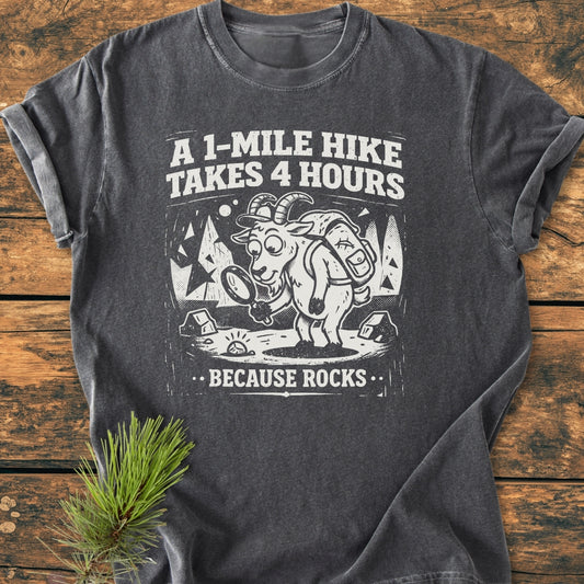 Because Rocks - Vintage Wash Tee featuring a cartoon goat with a backpack and magnifying glass against a forest, humorously captioned. Laid on a rustic surface with a pine sprig beside it.