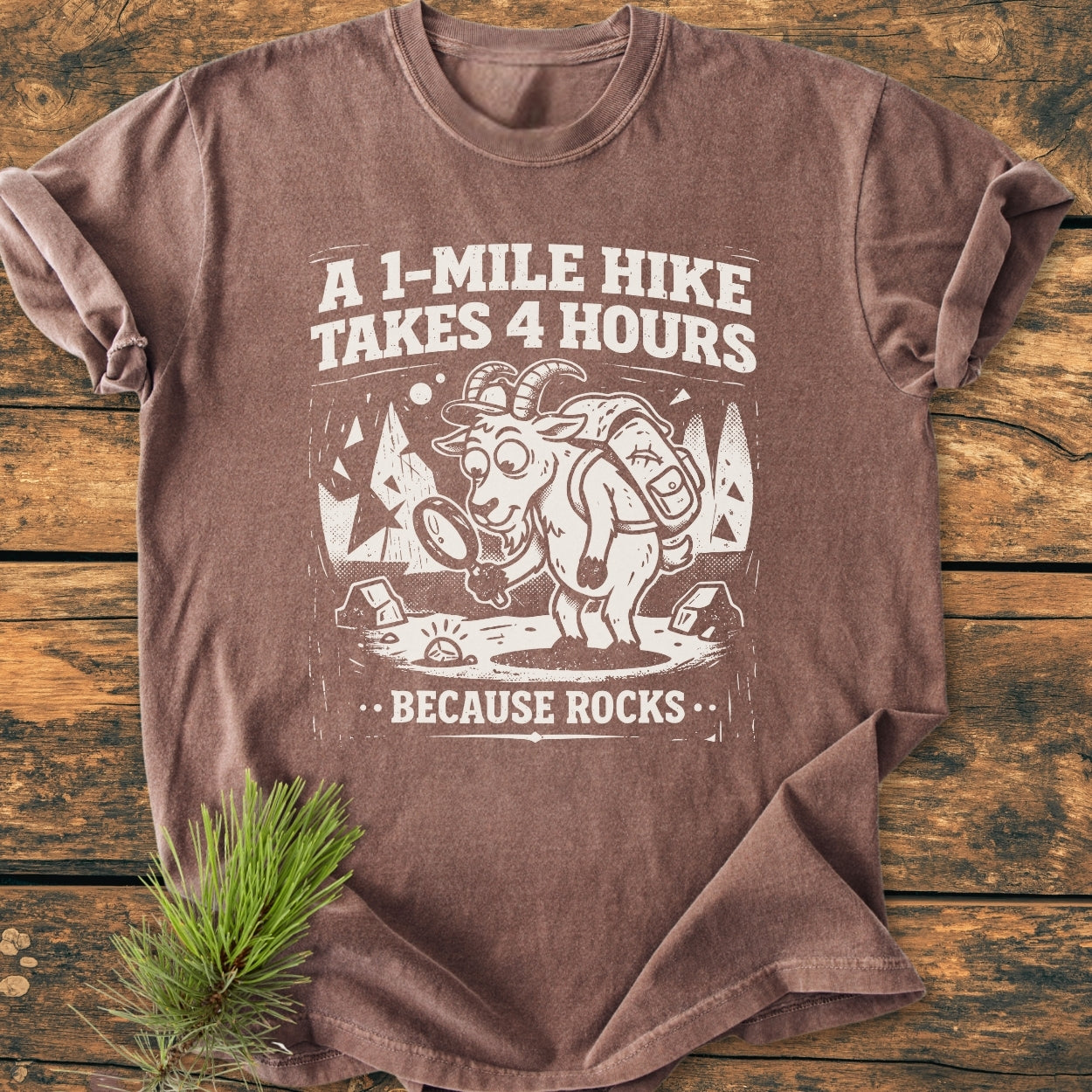 The Because Rocks - Vintage Wash Tee features a cartoon mountain goat with a backpack, magnifying a rock, and humorous text. It's laid on wood with a pine sprig beside it.