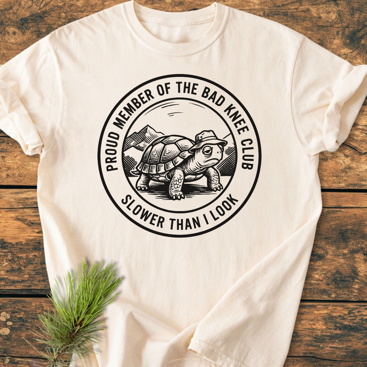 Bad Knee Club - Vintage Wash Tee features a turtle graphic with a mountain backdrop and humorous text. Laid on wood, accompanied by a pine sprig, highlighting its relaxed fit and durable construction.