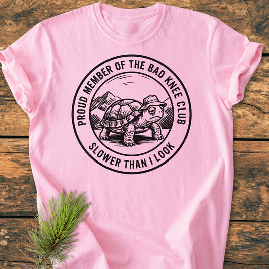 Bad Knee Club - Vintage Wash Tee: Light-pink T-shirt featuring a tortoise graphic with text, “PROUD MEMBER OF THE BAD KNEE CLUB” and “SLOWER THAN I LOOK,” laid on wood with pine sprig beside it.