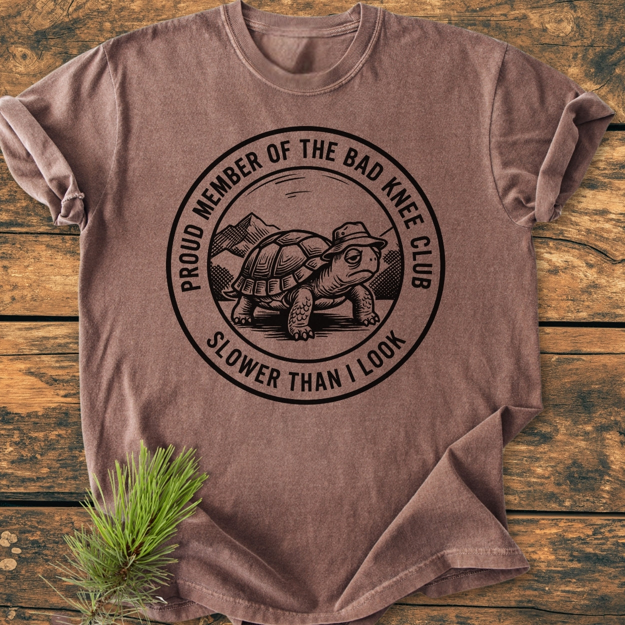 Bad Knee Club - Vintage Wash Tee featuring a turtle logo with humorous text, displayed on a wooden surface with a pine sprig. Made from soft, garment-dyed, ring-spun cotton, offering a relaxed fit.