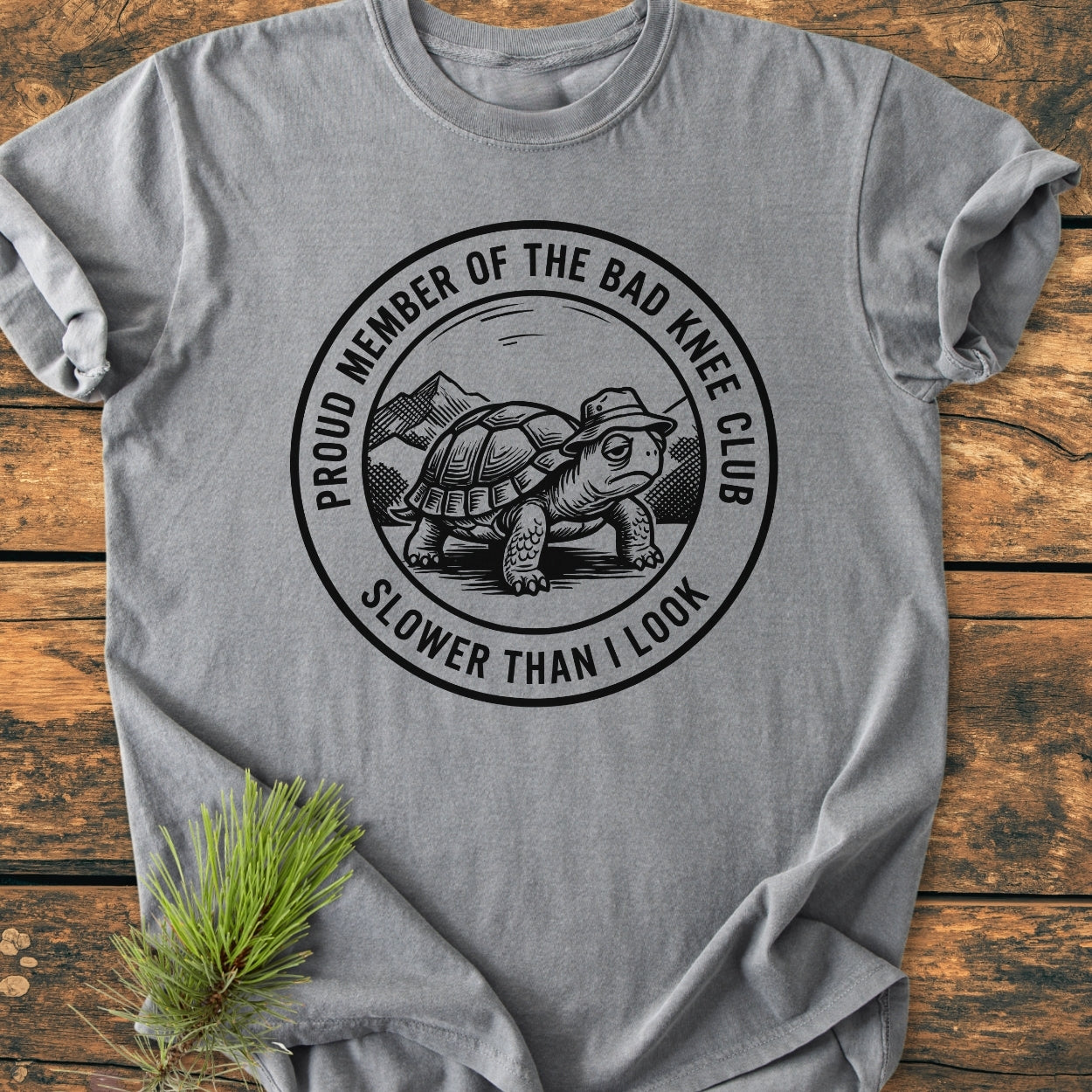 Bad Knee Club - Vintage Wash Tee features a turtle graphic with a bucket hat, set in a mountain scene, on a heather-gray tee, laid flat with a pine sprig accent.