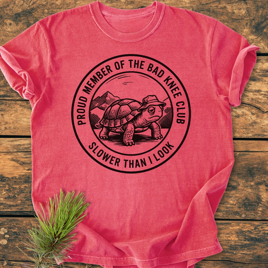 Bad Knee Club - Vintage Wash Tee features a black graphic of a turtle with a hat in a mountain scene, text reads “Proud Member of the Bad Knee Club” and “Slower Than I Look.”