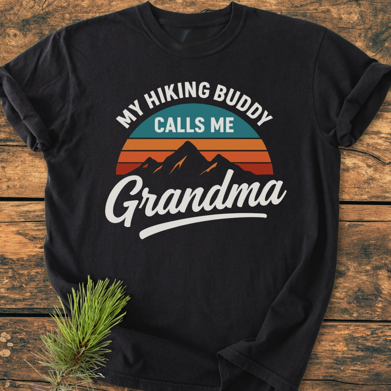 Hiking Buddy Grandma - Vintage Wash Tee features a retro mountain graphic with sunset stripes on a black T-shirt, laid on wood with a spruce branch, highlighting comfort and durability.