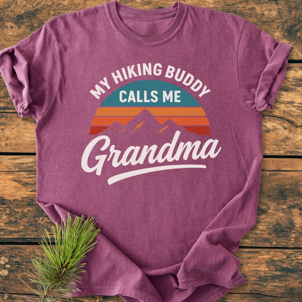Hiking Buddy Grandma - Vintage Wash Tee: A mauve T-shirt featuring a retro sunset and mountain graphic with the slogan. Laid flat on wood, adorned with a pine sprig accent.