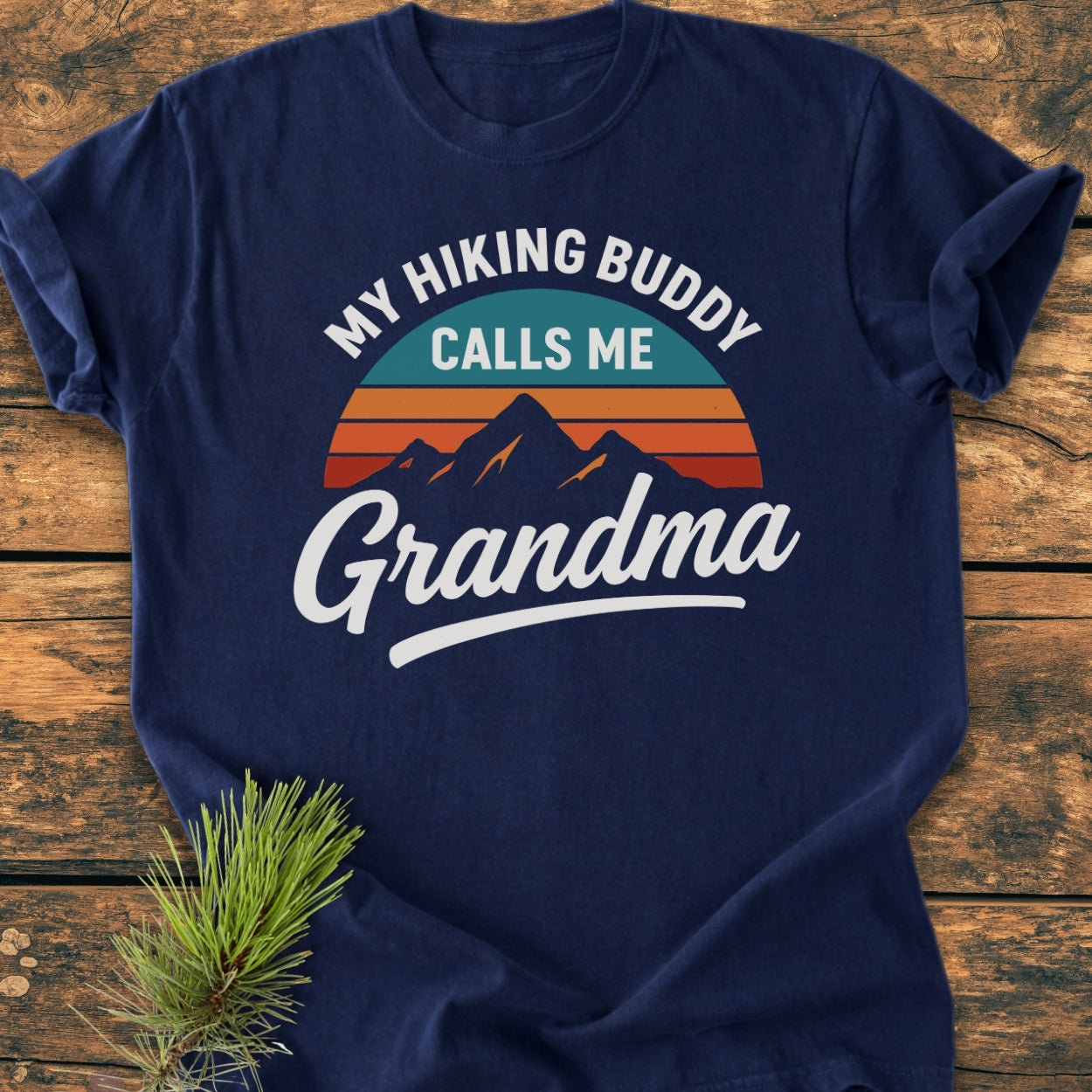 The Hiking Buddy Grandma - Vintage Wash Tee features a sunset/mountain graphic and slogan on a navy-blue T-shirt, laid on rustic wood with a pine sprig accent, showcasing its relaxed, cozy fit.