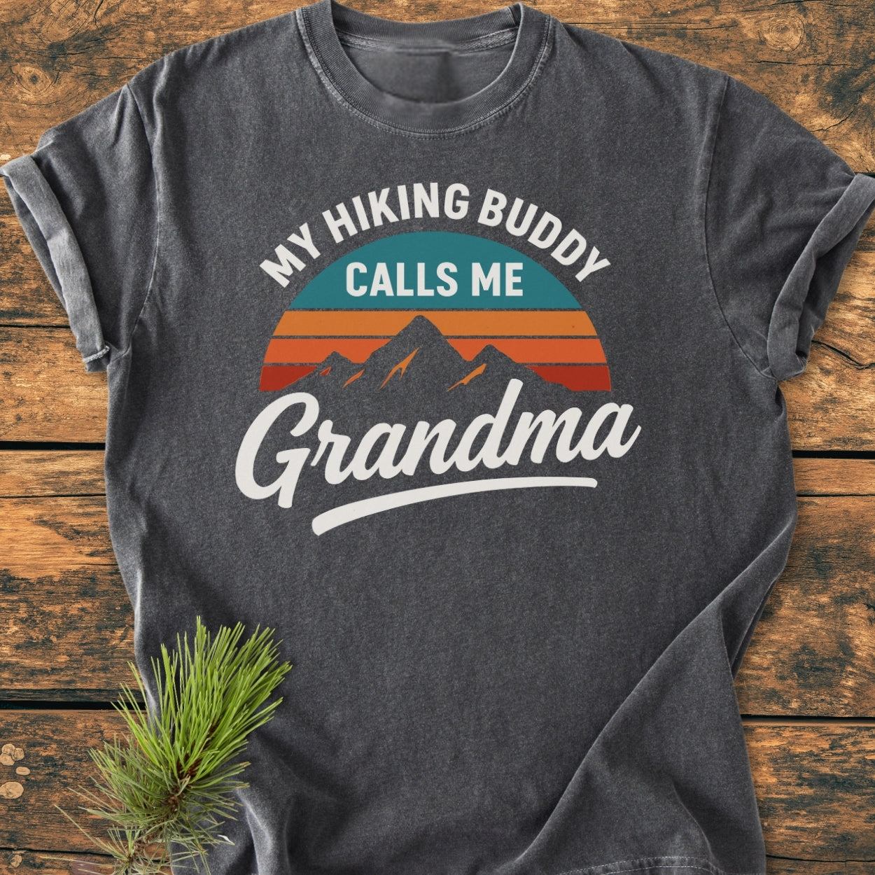 Hiking Buddy Grandma - Vintage Wash Tee in dark-heather gray, featuring a stylized sunrise mountain graphic. Laid on wood, it embodies comfort with a relaxed fit and durable, soft-washed cotton.