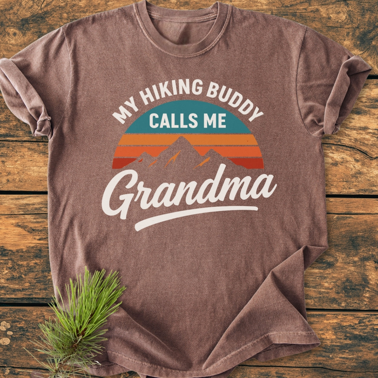 Hiking Buddy Grandma - Vintage Wash Tee: Heather-brown, relaxed-fit T-shirt with a retro mountain and sunset graphic, featuring MY HIKING BUDDY CALLS ME Grandma text, laid on a wooden surface with an evergreen sprig.
