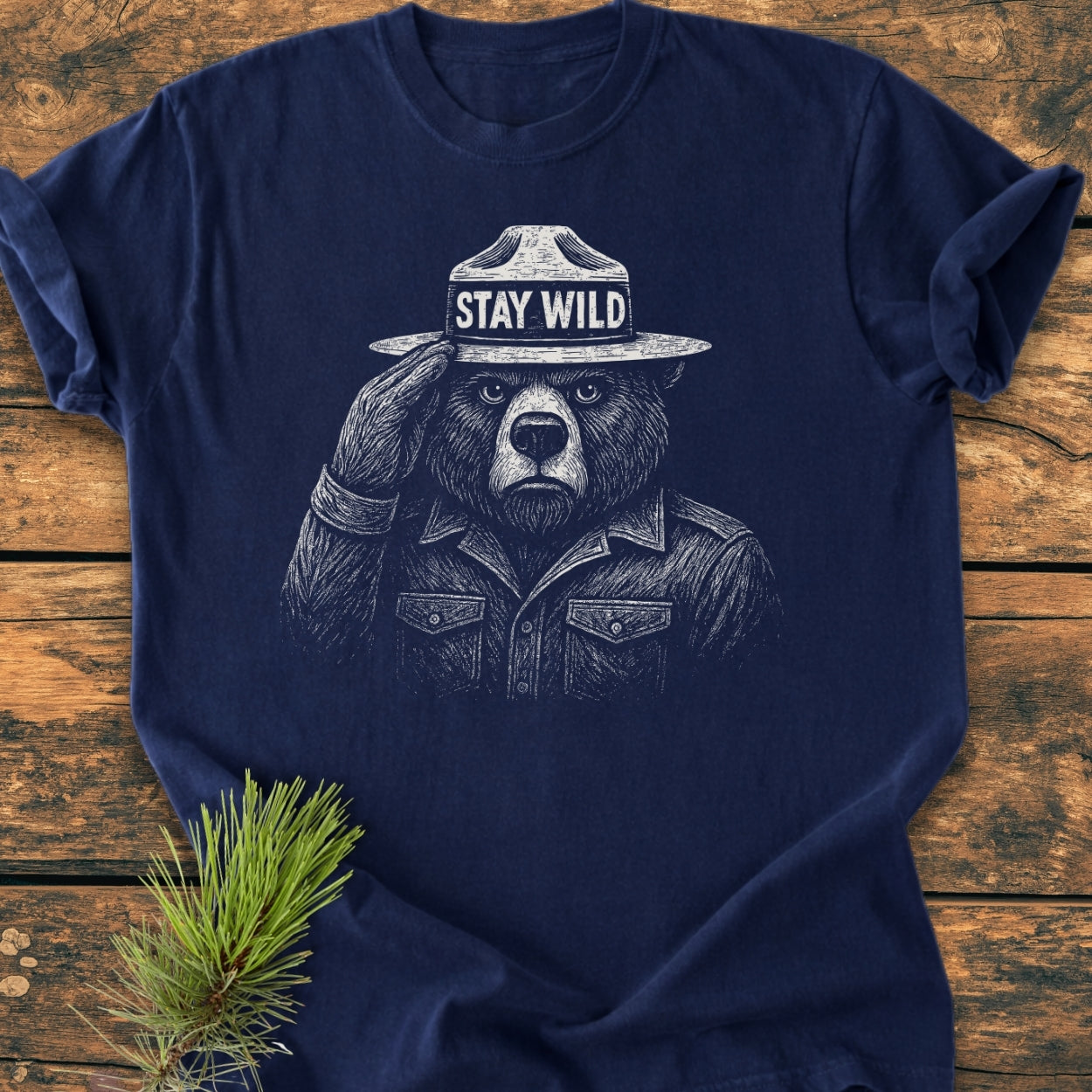 Stay Wild - Vintage Wash Tee features a bear graphic with a ranger hat saluting. Laid flat on rustic wood, this navy-blue, 100% ring-spun cotton tee offers durability and a relaxed fit.