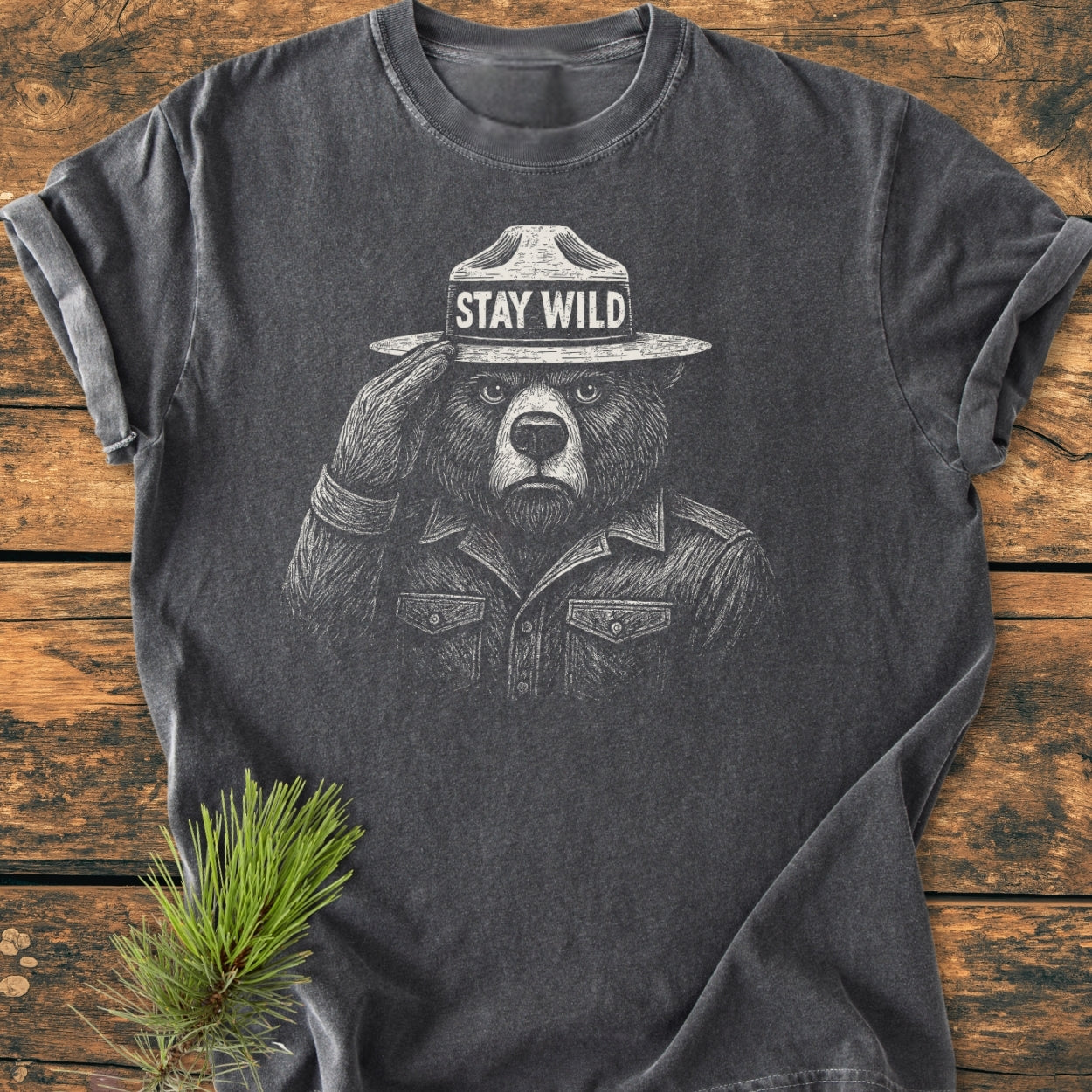 Stay Wild - Vintage Wash Tee features a bear in a ranger hat and button-front shirt, screen-printed in white on a charcoal-gray T-shirt, laid on rustic wood with a pine sprig beside it.