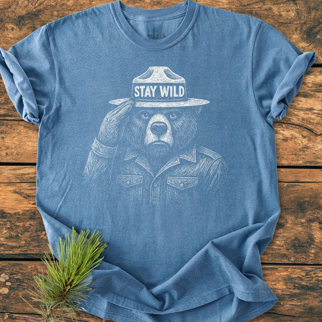 Stay Wild - Vintage Wash Tee featuring a sketched bear in a park-ranger hat with STAY WILD text, laid flat on a wooden surface with a pine sprig for an outdoorsy touch.
