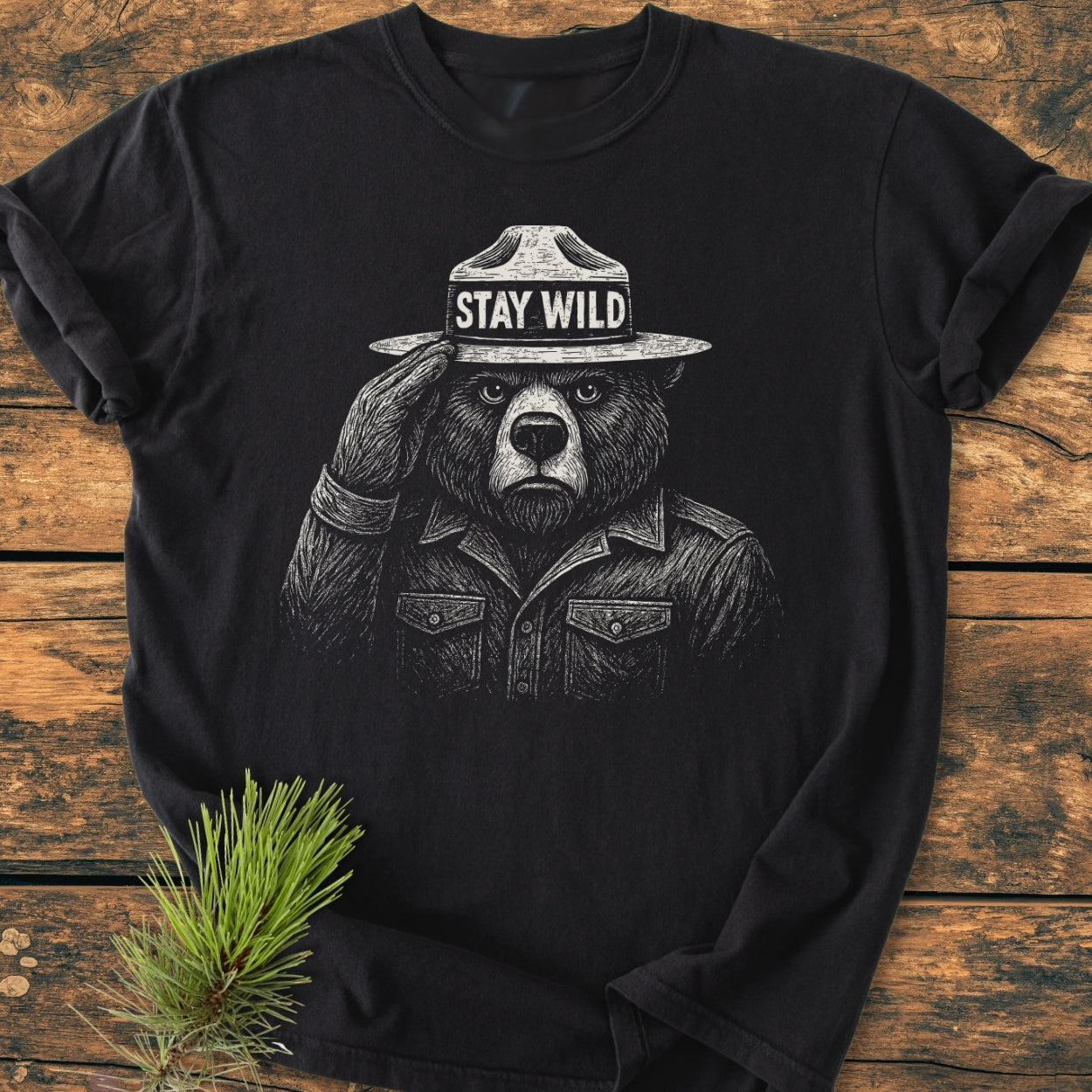 Stay Wild - Vintage Wash Tee featuring a white line-art bear in a ranger hat with STAY WILD, saluting. Laid on a wooden surface, showcasing its relaxed fit and durable stitching.