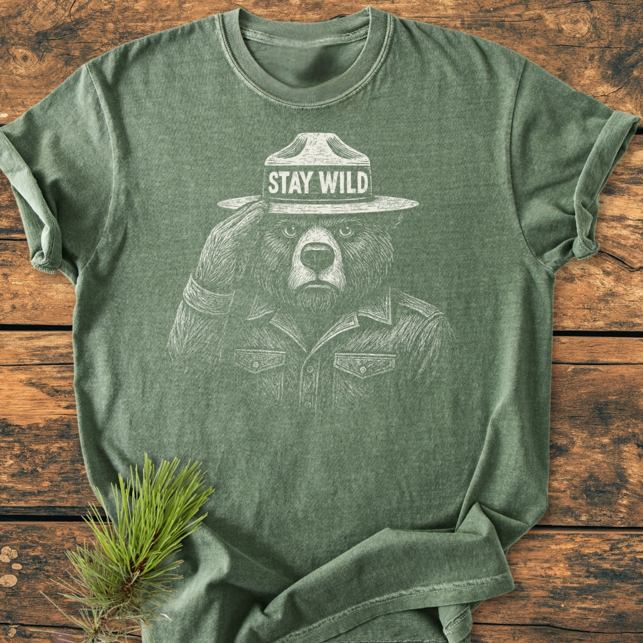 Stay Wild - Vintage Wash Tee features a bear illustration with a ranger hat on a heather-green T-shirt, laid on a wooden tabletop, with a pine sprig beside it, highlighting its rustic charm.