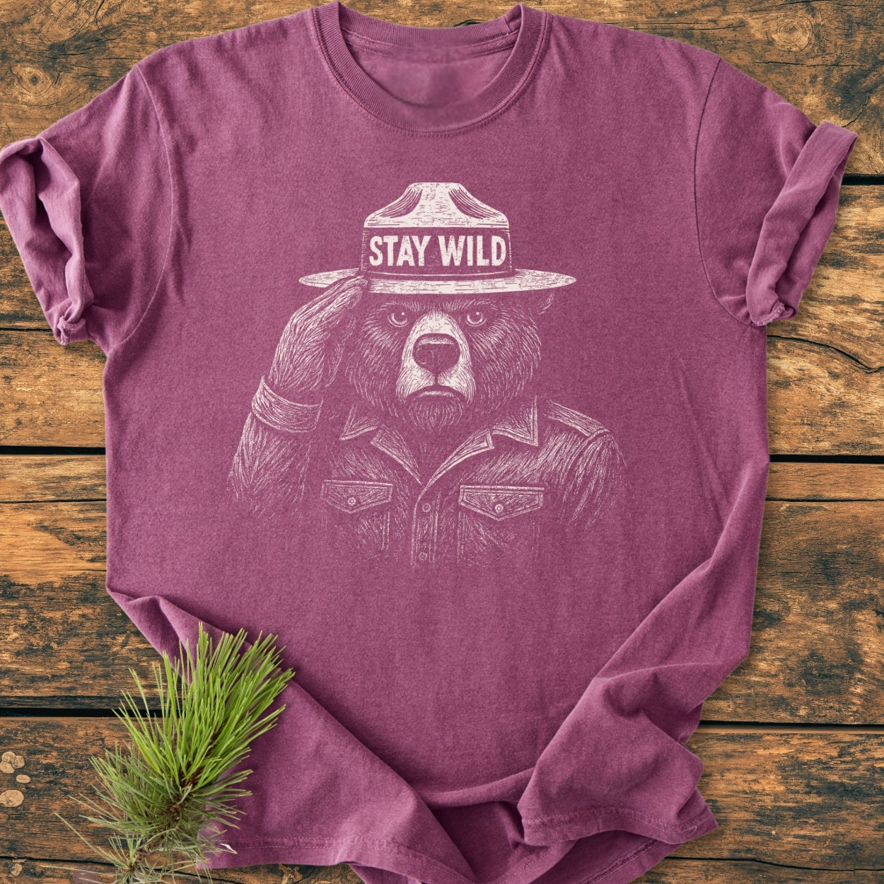 Stay Wild - Vintage Wash Tee featuring a bear dressed as a ranger, printed on a heather-purple long-sleeve T-shirt, laid on a wooden tabletop with a pine branch beside it.