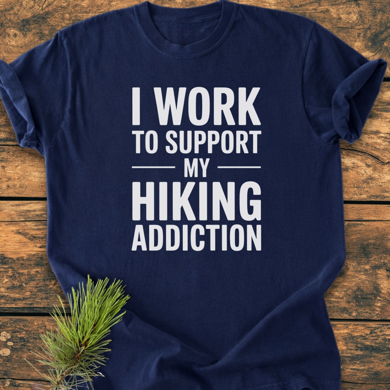 Hiking Addiction - Vintage Wash Tee: Navy-blue T-shirt with bold white text promoting hiking. Laid flat on wood, complemented by a pine branch, highlighting its outdoor theme and relaxed fit.
