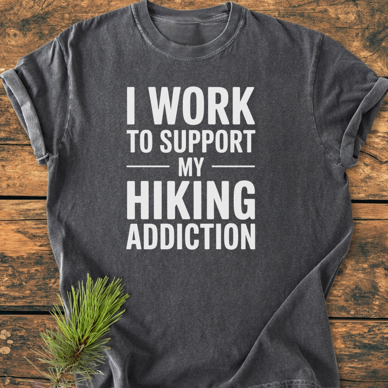 Hiking Addiction - Vintage Wash Tee displayed on a wooden surface, featuring bold white lettering. The charcoal-gray shirt showcases a relaxed fit, emphasizing its soft-washed, durable, tubular design ideal for hiking enthusiasts.