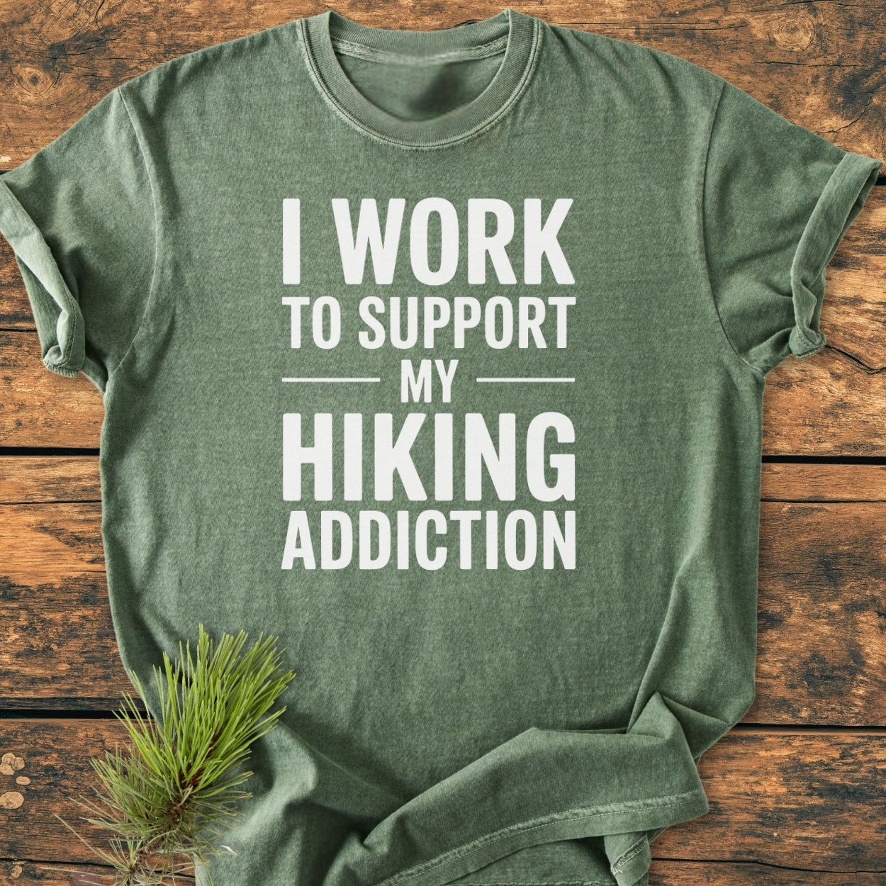 Heather-green Hiking Addiction - Vintage Wash Tee with white slogan on front, laid flat on wooden surface with pine sprig, showcasing its soft, relaxed fit and durable, ring-spun cotton fabric.