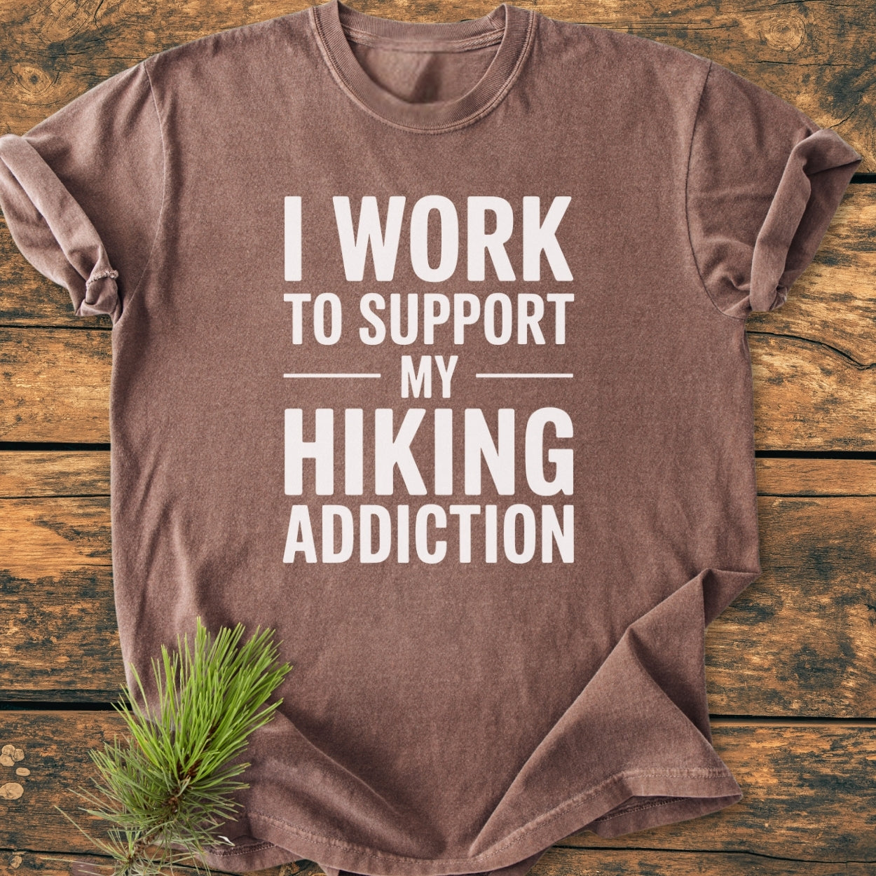 Hiking Addiction - Vintage Wash Tee, displayed folded on a wooden surface, features bold white text promoting a love for hiking. A pine branch accentuates the soft, relaxed fit of this durable, cozy shirt.