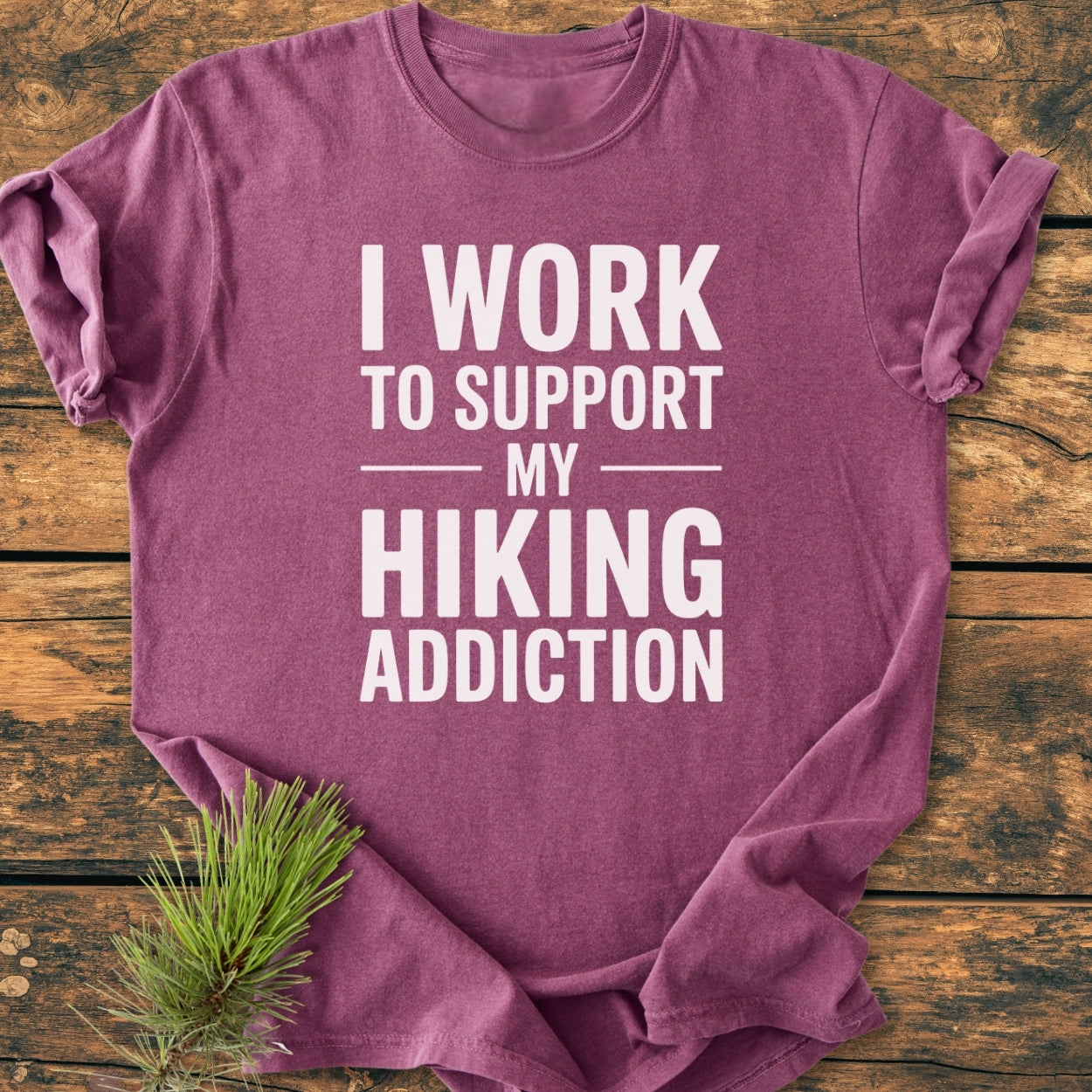 Hiking Addiction - Vintage Wash Tee laid flat, displaying bold white slogan about supporting a hiking addiction. Beside it, a small pine sprig emphasizes the outdoor theme.