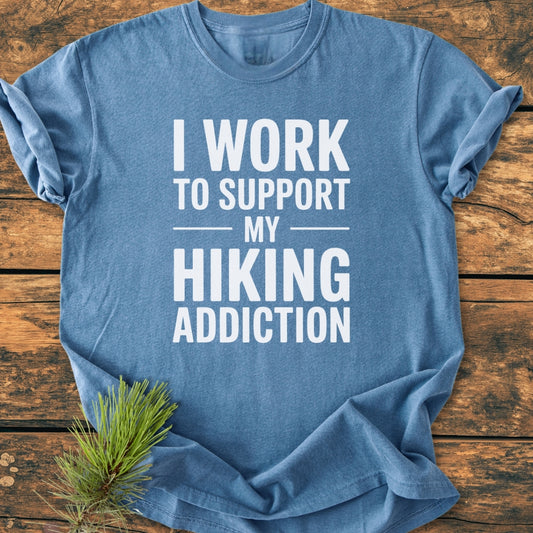 Hiking Addiction - Vintage Wash Tee displayed on wood surface, featuring bold white slogan. Includes sprig of pine. Made from soft, pre-shrunk 100% ring-spun cotton with a relaxed fit.