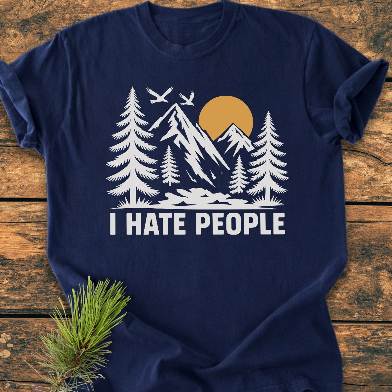 I Hate People - Vintage Wash Tee features a nature scene with mountains, sun, and birds. Relaxed fit, navy-blue, 100% ring-spun cotton, laid on rustic wood with pine sprig.