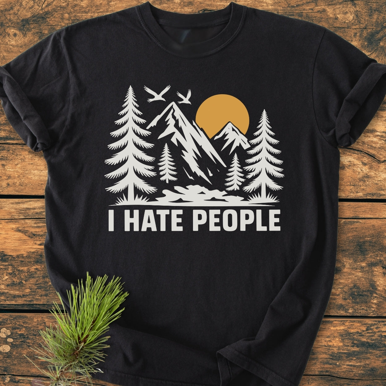 I Hate People - Vintage Wash Tee features a black tee with a white outdoor scene and bold text, laid flat with a pine sprig beside, showcasing its relaxed fit and durable stitching.