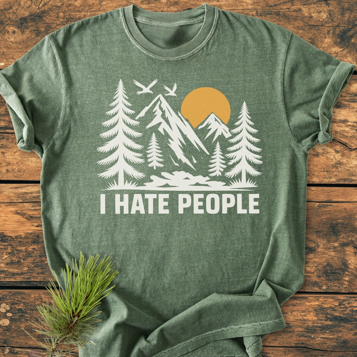 Forest-green I Hate People - Vintage Wash Tee featuring a stylized outdoor scene with trees and mountains, laid on rustic wood. Pine sprig beside the shirt highlights its nature-inspired theme.