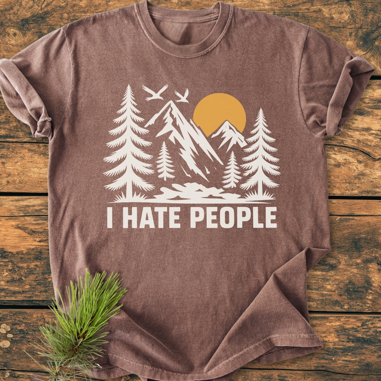 I Hate People - Vintage Wash Tee: Brown short-sleeved T-shirt with a stylized outdoor scene and bold text, laid flat on a wooden surface with a pine branch beside it.