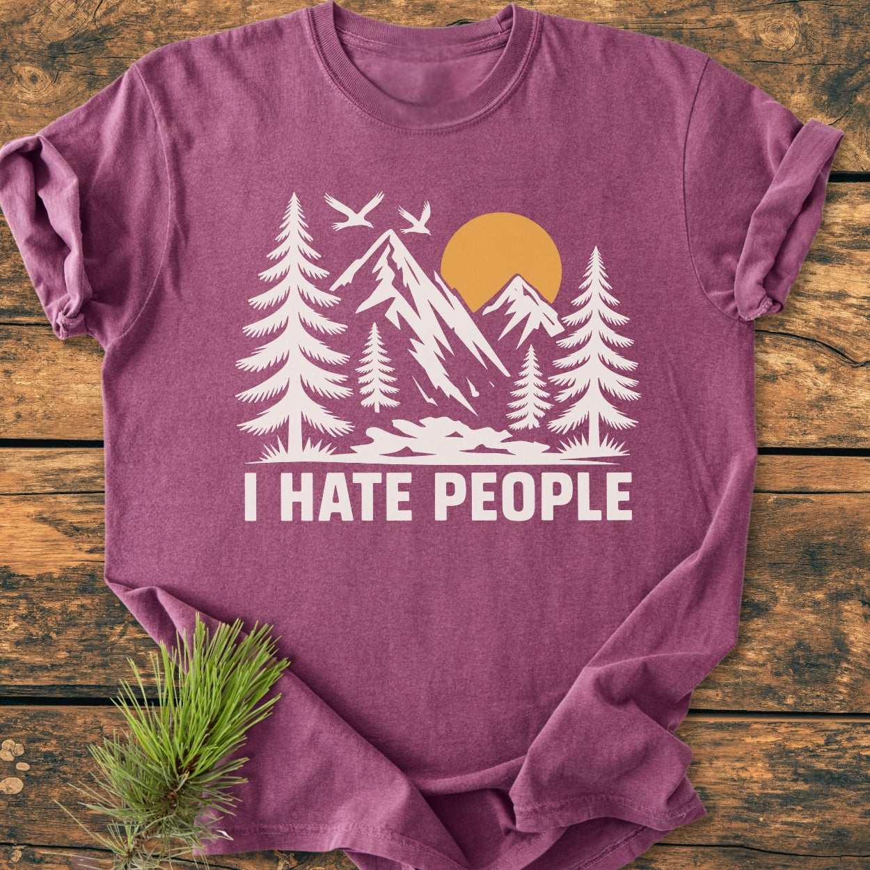 Heather-purple I Hate People - Vintage Wash Tee with a nature scene graphic, featuring pine trees, mountains, sun, and birds, laid flat on wood. Includes a pine branch accentuating the outdoorsy theme.