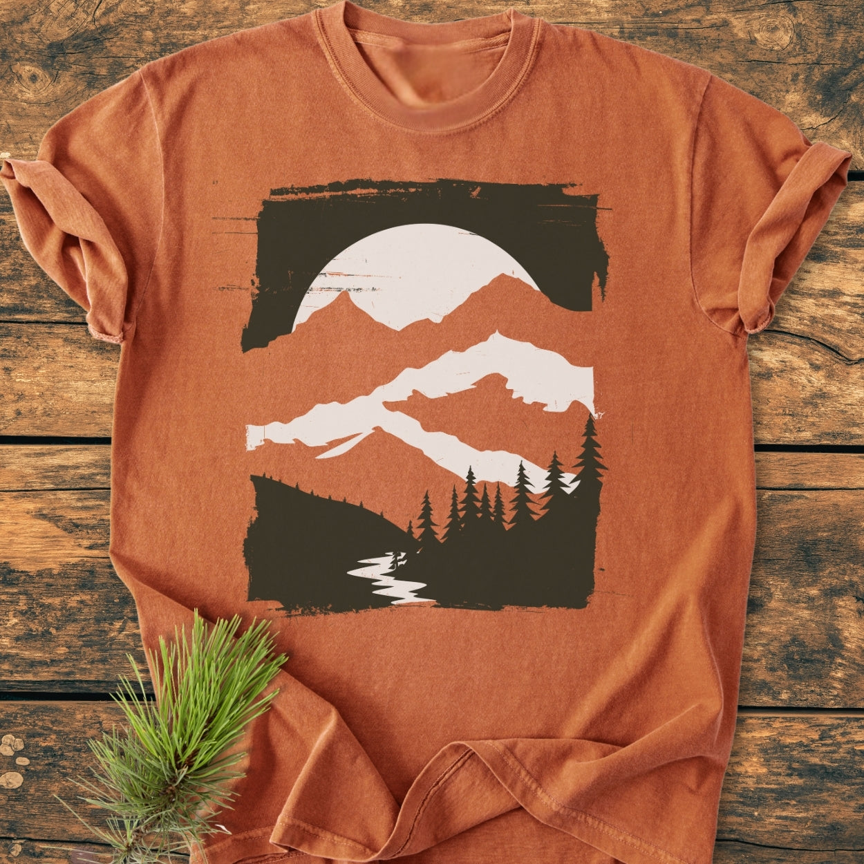 Minimalist Mountains - Vintage Wash Tee featuring a stylized outdoor scene with a sun, mountains, trees, and a river. The rust-orange shirt lies on wood, accented by a pine sprig.