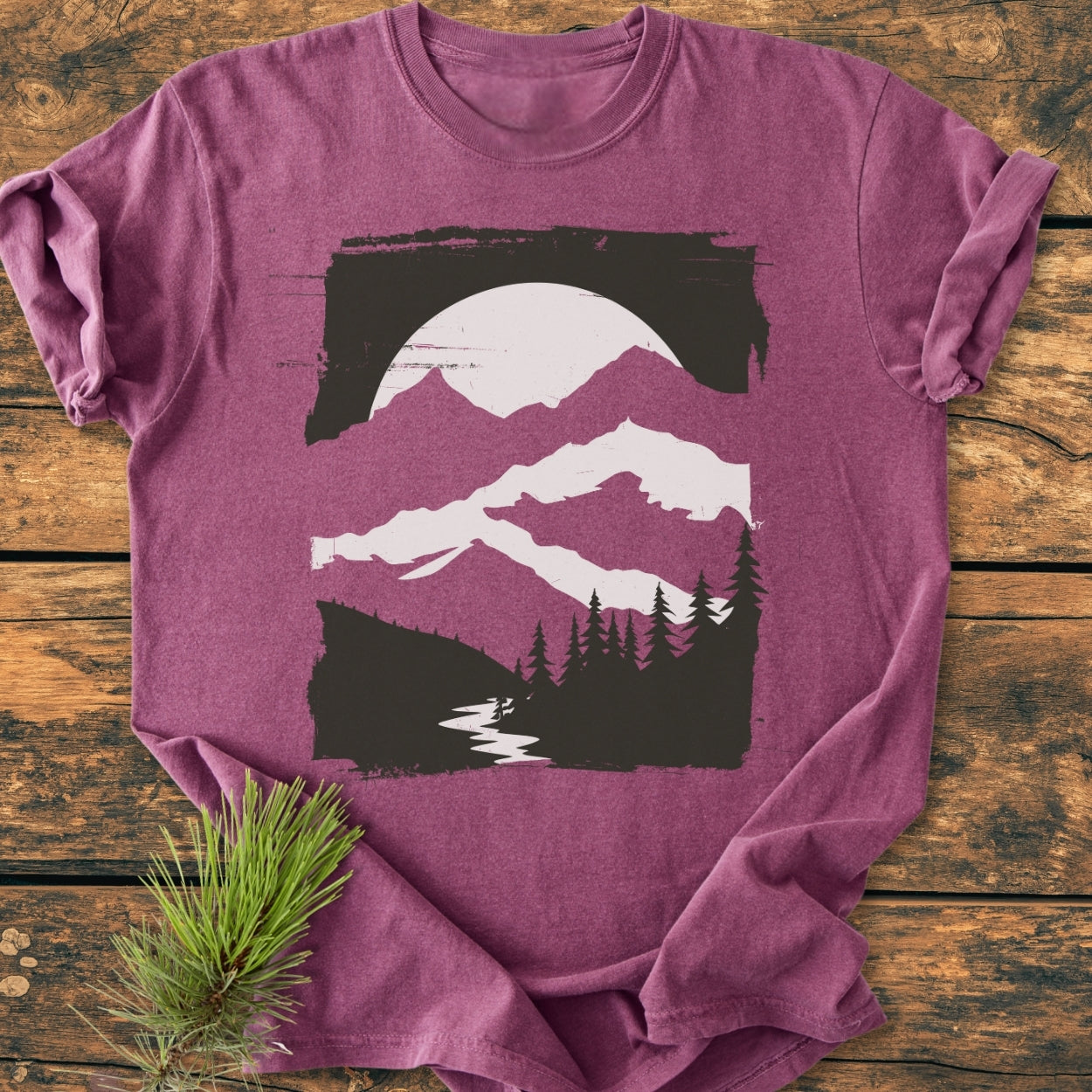 Minimalist Mountains - Vintage Wash Tee: A heathered plum-purple T-shirt with a black and white graphic of mountains, sun, pine trees, and a stream, laid flat on a wooden surface with a pine sprig beside it.