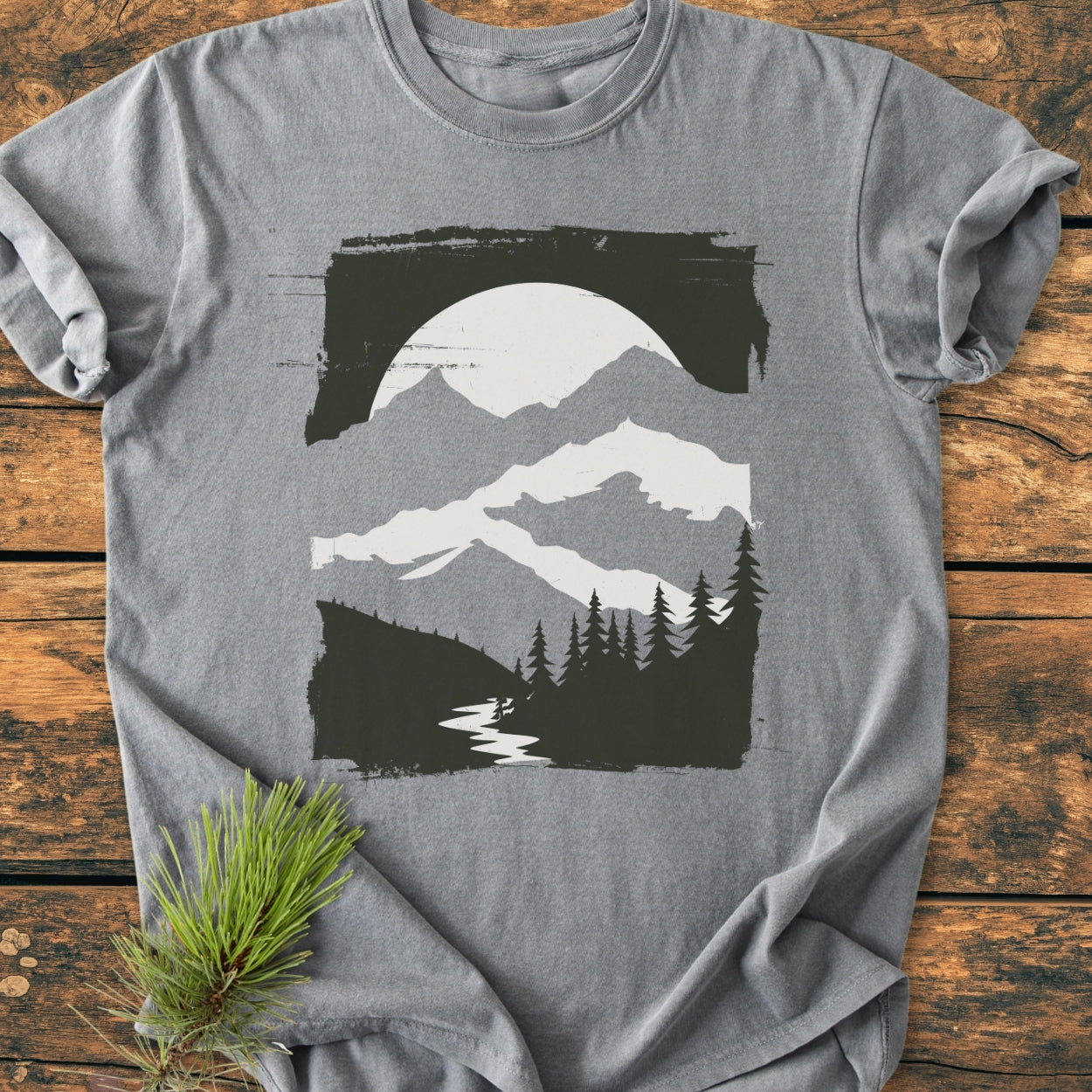 Minimalist Mountains - Vintage Wash Tee on rustic wood features a stylized landscape with pine tree silhouettes, a winding river, snow-capped mountains, and a sun or moon, highlighting its relaxed, cozy fit.