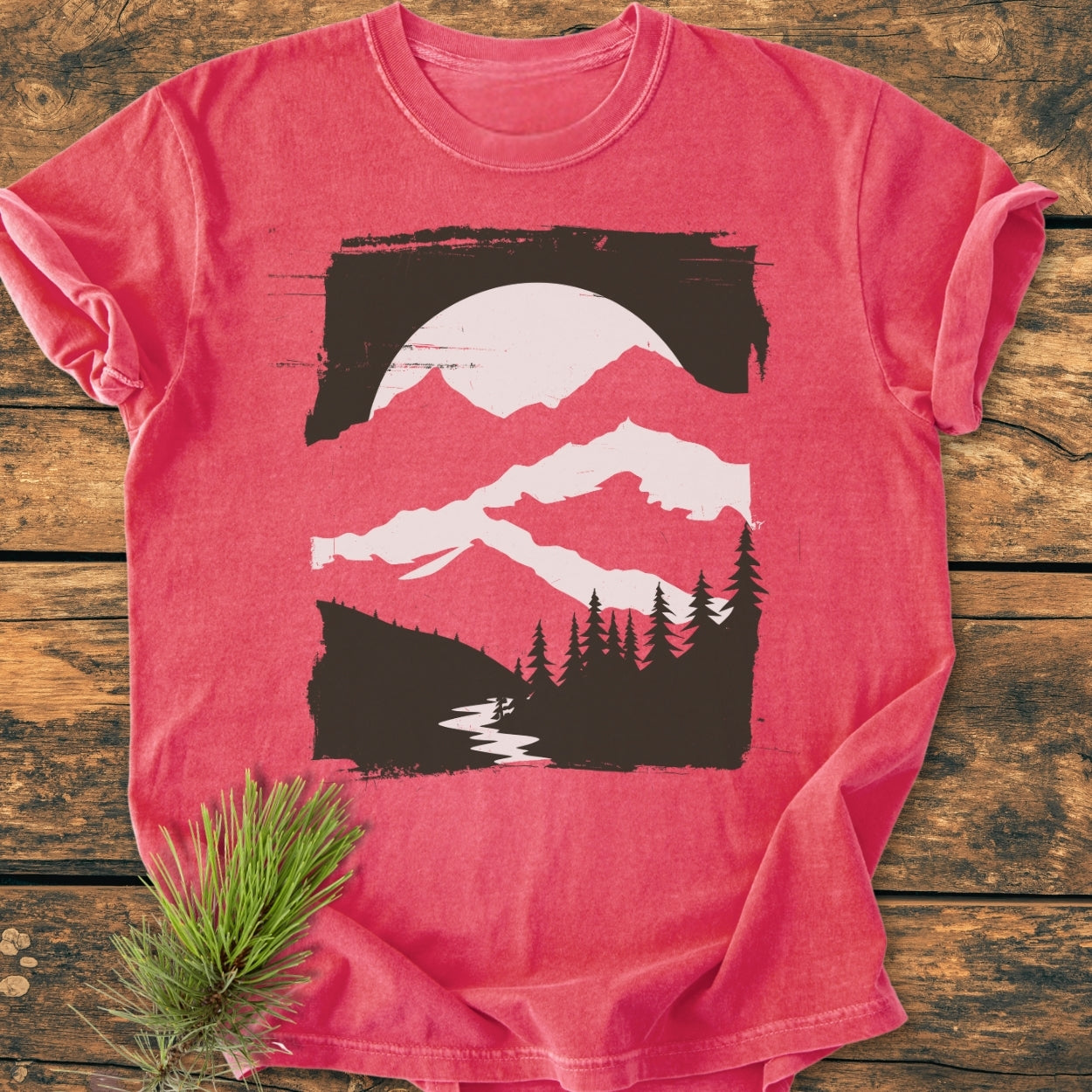 Minimalist Mountains - Vintage Wash Tee features a nature scene with mountains, sun/moon, and trees on a red crew-neck T-shirt, laid on rustic wooden planks, with a pine sprig accent.