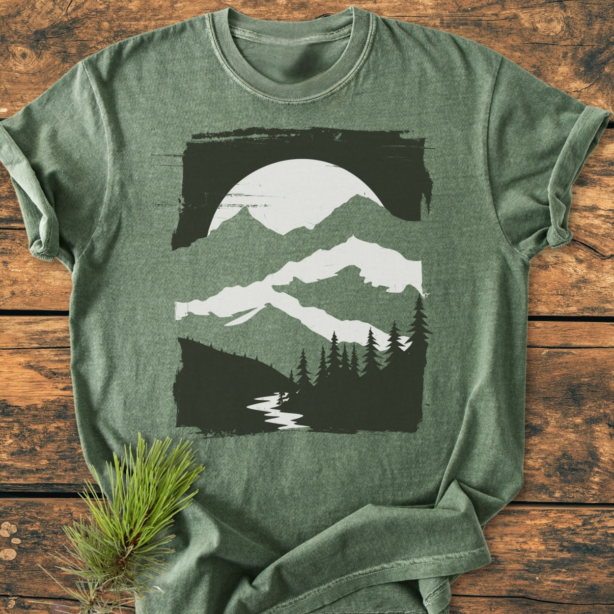 Minimalist Mountains - Vintage Wash Tee in heather-green, featuring a black and off-white nature scene with mountains, sun/moon, pine trees, and river. Displayed on rustic wood with pine sprig accent.