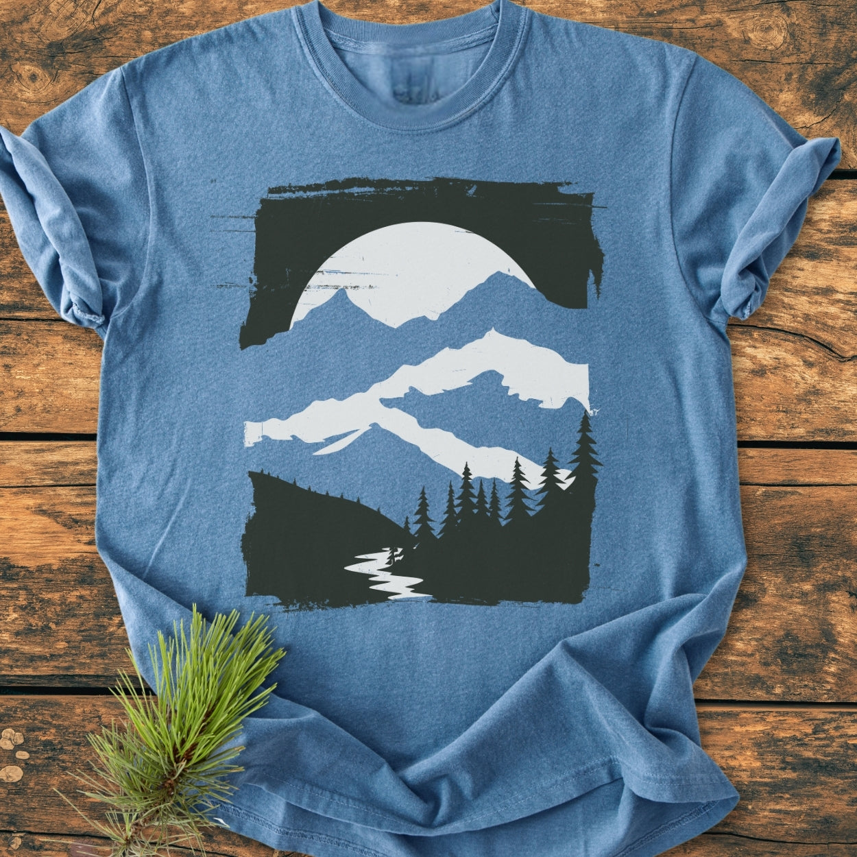 Minimalist Mountains - Vintage Wash Tee features a stylized mountain landscape with snow-capped peaks, evergreen trees, and a river, framed by a brush-stroke border, laid on wooden surface.